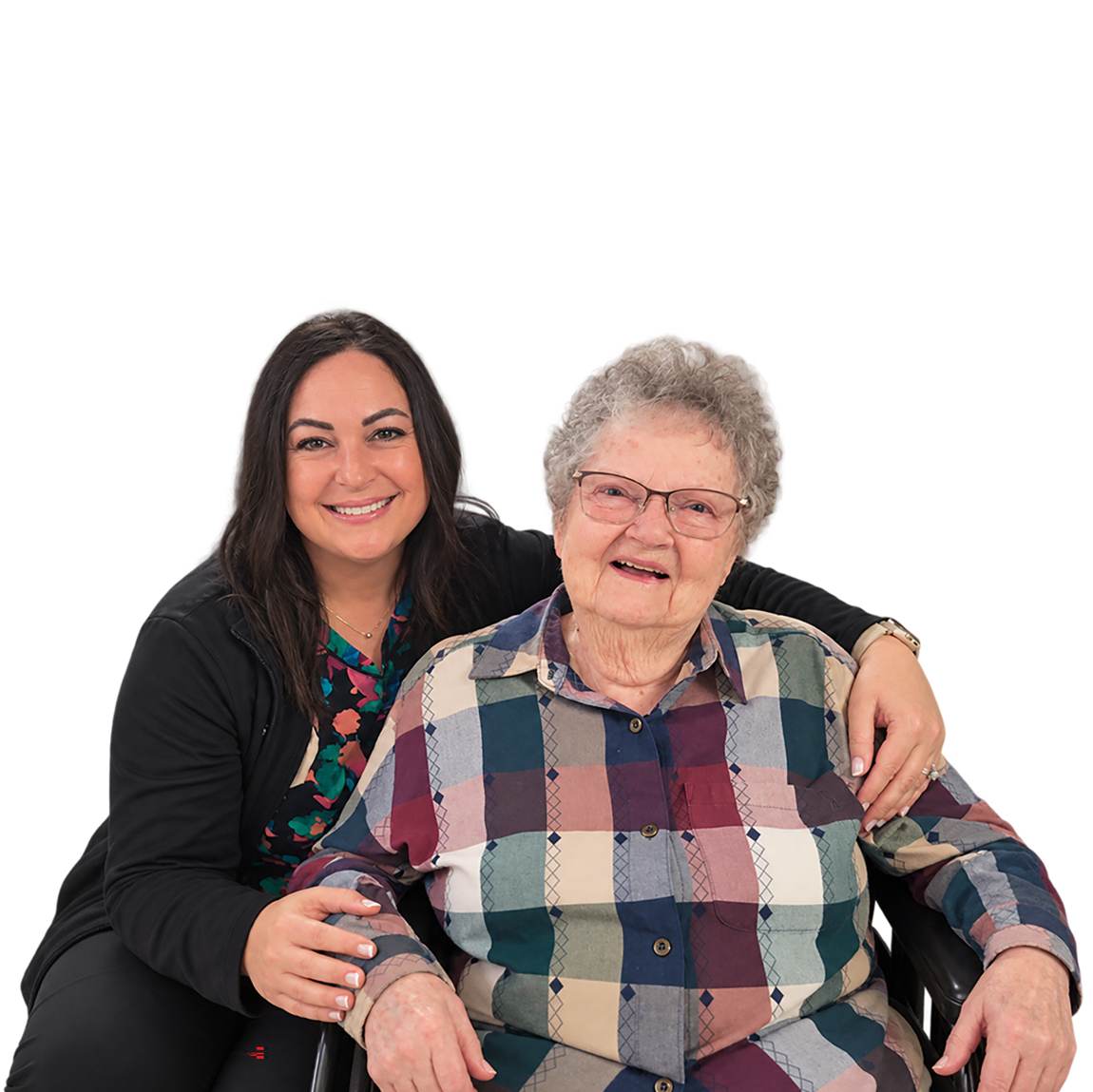 Two smiling women sit close together: a younger caregiver with dark hair embraces an elderly woman in a plaid shirt, conveying warmth and companionship.
